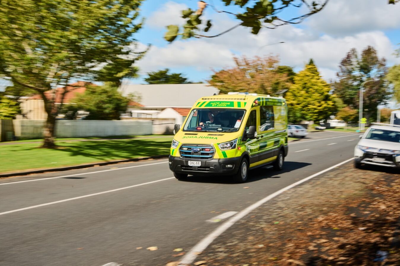 Hato Hone St John's new electric emergency ambulance driving on a road past a green tree in New Zealand